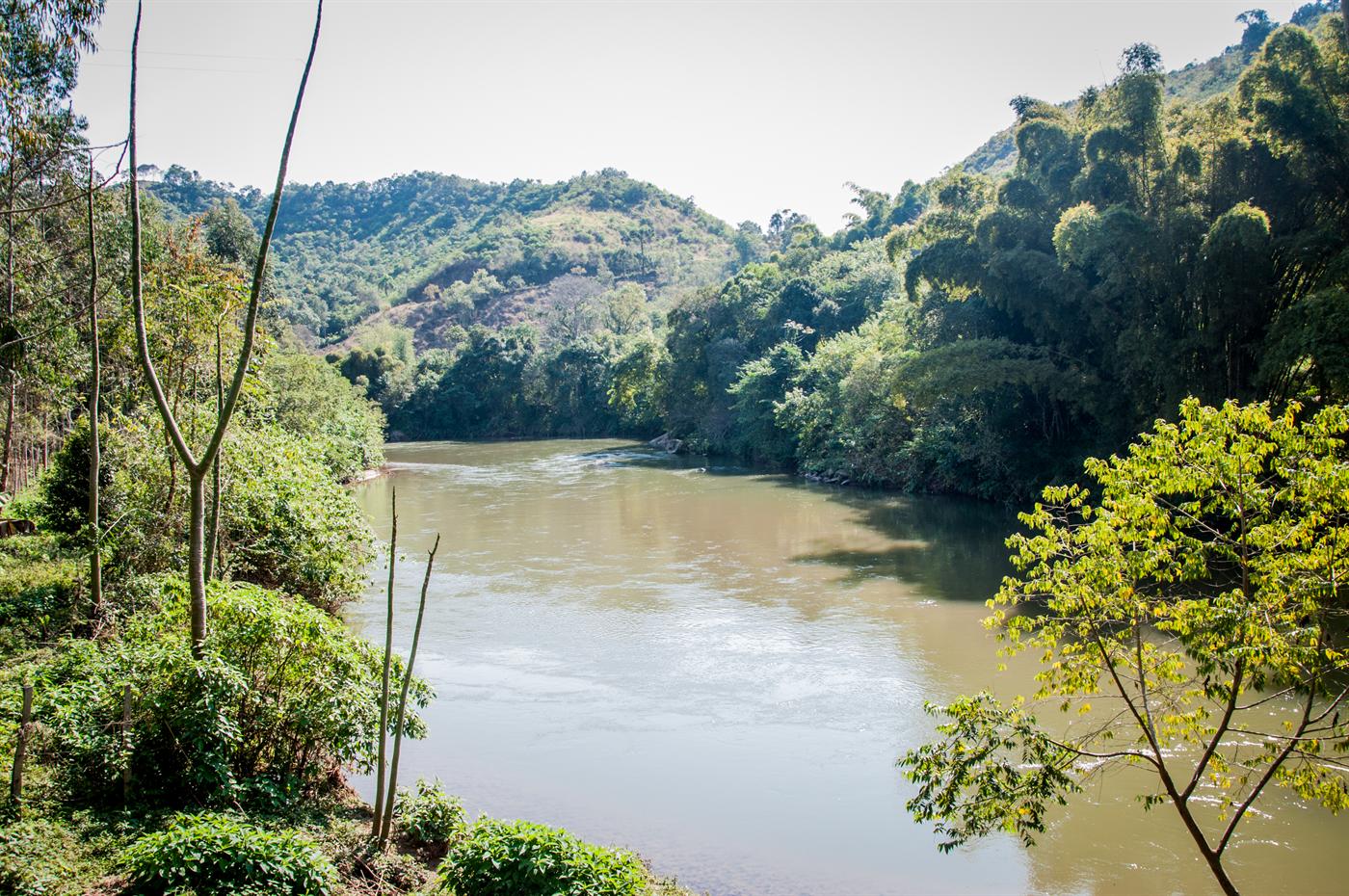 Rio visto da margem, entre árvores, com morros ao fundo